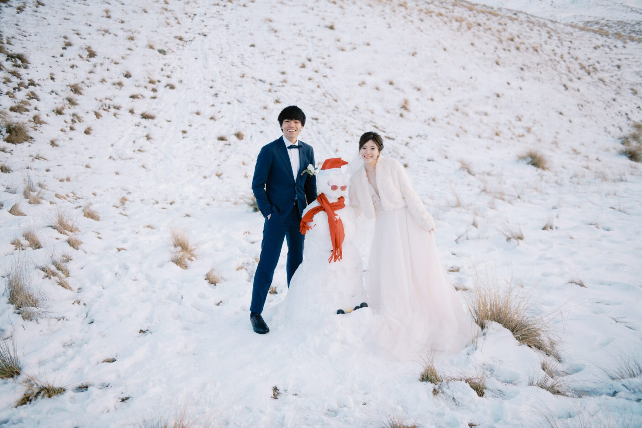 Queenstown Tekapo New Zealand Heli Wedding Elopement Pre-Wedding Shoot Photographer クイーンズタウン　テカポ　ニュージーランド　エロープメント 前撮り　フォトウェディング　結婚式 | A couple in wedding attire posing with a snowman in a snowy Tekapo landscape during their pre-wedding shoot.